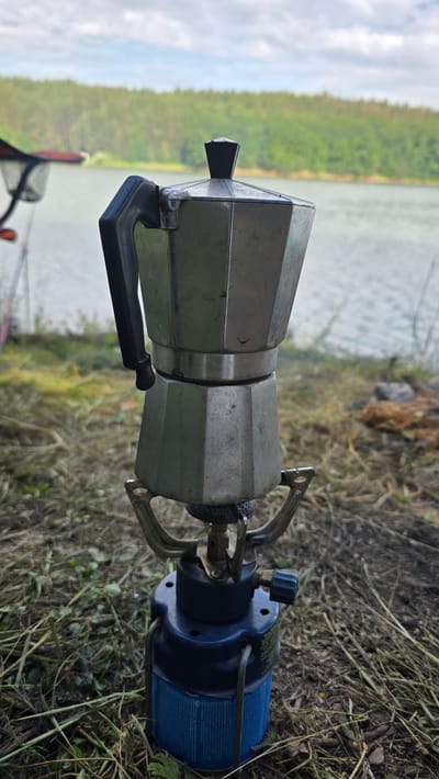 A stovetop espresso maker, likely aluminum, sits atop a blue portable camping stove with a propane canister. The espresso maker has a black handle and a faceted body, with visible signs of use. It is positioned outdoors on dry grass and dirt. In the blurred background, a body of water is visible, with a dense, green forest lining the opposite shore under a cloudy sky. The scene appears to be in a natural setting, possibly near Dolni Pasarel, Bulgaria, given the vegetation and water.