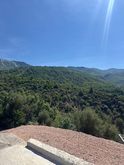 The image shows a scenic view of rolling green hills and mountains under a clear blue sky with a bright sun glare in the upper right corner. In the foreground, there is a concrete structure with a reddish gravel surface next to it. The hills in the midground and background are densely covered with lush green trees, creating a textured landscape. The top of one of the further mountains has some bare patches and what appears to be man-made structures, possibly telecommunication towers. The scene depicts a natural, mountainous environment.