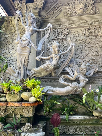 The image shows a wall in Kedewatan, Indonesia, adorned with sculptures. Three stone-colored figures of dancing women are positioned against a backdrop of stone carvings depicting a scene with multiple figures. The figures are wearing ornamented cloths, jewels and elaborate headdresses, and are frozen in fluid motions of dance. The background wall is decorated with carvings of figures and foliage. The base of the wall is constructed from stacked stones. The side of the display is adorned with assorted plants and stones.
