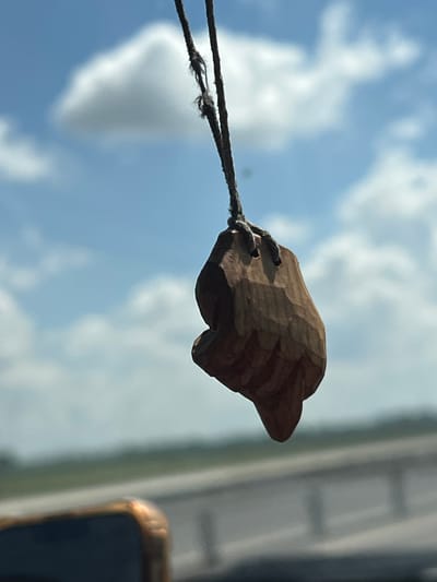 A close-up, horizontal shot captures a small, carved wooden object resembling a hand or fist, suspended by a frayed rope against a backdrop of a partly cloudy blue sky. The wooden carving is dark brown and appears textured with visible tool marks. It is attached to two strands of dark, weathered rope. The rope extends to the left and appears to be attached to something out of frame. The sky is bright, suggesting daylight, with fluffy white clouds scattered across the blue expanse. In the far right of the frame, a blurry vertical element with a golden-brown edge is visible, possibly the corner of a window or frame. The lighting and sky suggest it is daytime, likely a sunny or partly sunny day. There is no visible text. The setting appears to be outdoors with a view of the sky. The overall impression is simple and somewhat rustic, focusing on the natural textures of the wood and rope against the vastness of the sky.