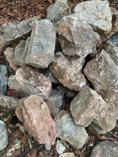 A pile of large, rough, gray and brown rocks lies on the ground, interspersed with dry leaves and some green foliage. The rocks are of various shapes and sizes, with many exhibiting sharp edges and irregular surfaces. Some rocks have lighter colored patches of white or pale gray, possibly indicating mineral deposits or weathering. The scene appears to be outdoors, with the ground covered in a layer of fallen leaves and dirt, suggesting an uncultivated or natural setting. There is a small orange object, possibly a piece of fruit or debris, visible among the leaves and rocks in the upper left portion of the image. The lighting suggests it is daytime. No people are visible in the image. The location of this scene in Olhão, Portugal is not provided in the image itself.