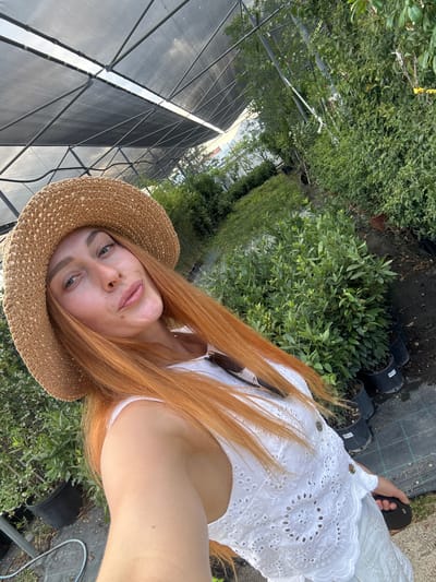 A young woman with long, auburn hair and a straw hat is posing for a selfie in a greenhouse. She is wearing a white eyelet top. The greenhouse is filled with rows of green plants in black pots. The roof of the greenhouse is made of a dark mesh material supported by metal beams. The ground is covered with gravel and black plastic sheeting. The scene appears to be outdoors, possibly in a nursery or garden center, in Radanovići, Montenegro.