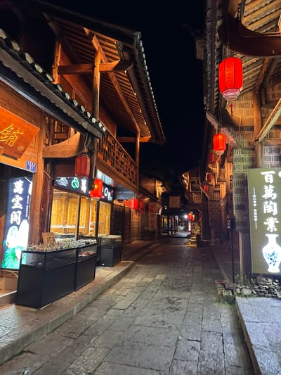 The image depicts a narrow street in the city of Zhuoshui, China, at night. The street is lined with traditional wooden buildings adorned with red lanterns, signs with Chinese characters, and product display cases. The street is paved with stone. The overall atmosphere is warm and inviting, illuminated by the lights from the buildings and lanterns. The image captures the architectural details of the buildings and the narrowness of the street. The OCR provides written words from the signs on the buildings.