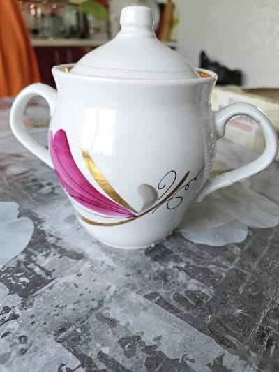 A white ceramic sugar bowl with pink and gold floral accents is centered on a patterned tabletop. The sugar bowl has two handles and a lid. The tabletop has a mottled grey and white design that appears textured. The background is blurred but suggests an indoor setting with some kitchen elements, including an orange fabric item and shelves with various objects. Crystals of what appears to be sugar are sprinkled on the handles and rim of the sugar bowl and on the tabletop around it.