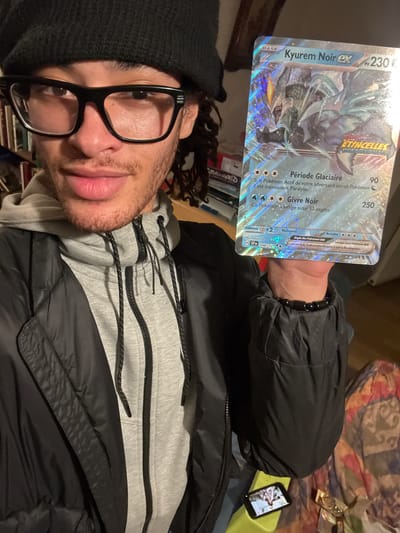 A person with dark curly hair, a black beanie, and black-rimmed glasses is holding a French Pokémon card of “Kyurem Noir EX” in front of them in an indoor setting. The card is silver with the Pokémon’s image and attack descriptions, the card number is visible as "048/115." The person is wearing a grey hooded sweatshirt under a black puffy jacket. A beaded bracelet is visible on their wrist. In the background is a shelf of books and a cell phone.