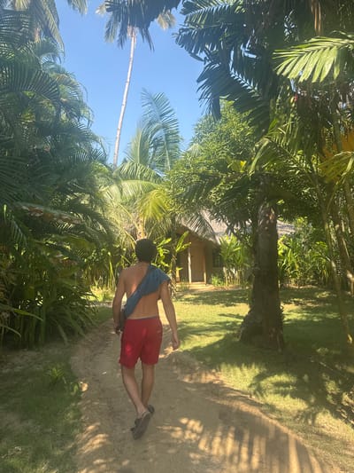 A partially-nude, medium-skinned male in red shorts walks on a tan dirt path in a tropical environment, carrying a blue bag over his shoulder, underneath a blue sky. Lush, green tropical foliage, including palm trees, surrounds the path. A small, tan-colored structure with a thatched roof is visible in the background. The image was taken in El Nido, Philippines.