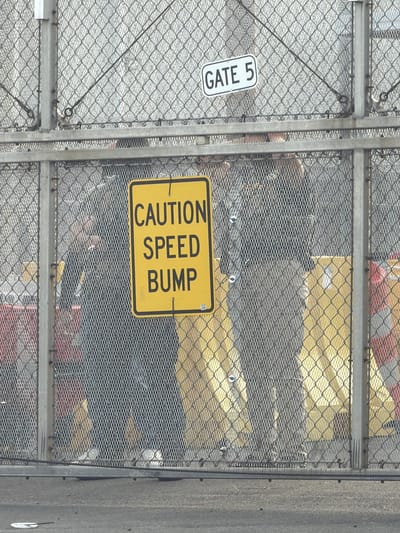 The image shows a chain-link fence with a "GATE 5" sign attached to the left side. A "CAUTION SPEED BUMP" sign is affixed to the fence's right side. Behind the fence, there are indistinct shapes, including hints of yellow, gray, red, and black, possibly representing objects or clothing. The location is the city of Newark, United States of America.