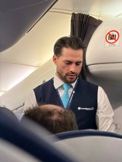 A man with dark hair and a beard is asleep in an airplane. He wears a white shirt, blue tie, and a dark blue vest with the Aeromexico logo. The airplane's interior is visible, including a sign prohibiting luggage in the overhead compartment. The cabin ceiling and a partially visible seated passenger are also present in the frame.