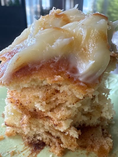 A close-up, overhead shot shows a piece of cake with a cinnamon sugar filling and a white glaze. The cake is light brown and has a crumbly texture. The cinnamon sugar filling is dark brown and appears to be layered within the cake. The white glaze is thick and glossy, dripping over the sides of the cake. The cake is served on a pale green plate. The background is blurred and out of focus, but appears to be a dark indoor setting with some light reflecting off surfaces. The lighting suggests it is daytime.