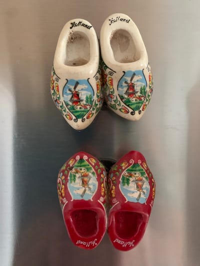 The image shows four miniature wooden clogs, which are traditional Dutch footwear. Two clogs are white and two are red, all adorned with painted scenes of windmills and floral patterns. The word "Holland" is written in script on the side of each clog. The clogs are arranged in two pairs, with the white clogs on the left and the red clogs on the right. They appear to be decorative items, possibly magnets or souvenirs, resting on a reflective, metallic surface. The lighting suggests the photo was taken indoors, with no specific time of day or weather conditions discernible. There are no people or any activity taking place in the image. The location context of Ivanava, Belarus, is external information and not visually evident from the image itself.