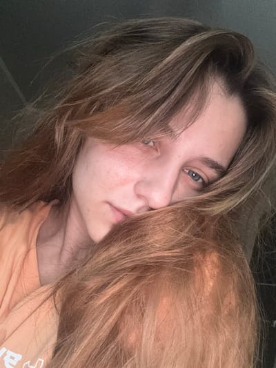 The image is a close-up selfie of a young person with light skin, blue eyes, and light brown hair. They are wearing an orange t-shirt. The person is looking towards the camera with a neutral expression, with their hair partially covering their face. The background is dark and out of focus, suggesting an indoor setting. The image does not contain any information that would indicate the location in the city of Vyksa, Russia.