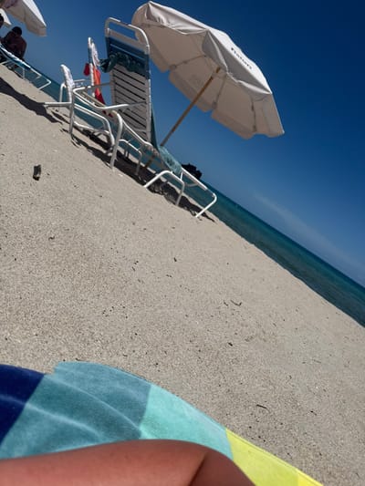 A low-angle view captures a sunny beach scene in Hallandale Beach, United States. In the immediate foreground, the lower leg of an individual, resting on a horizontally striped towel (blue, light blue, green, and yellow), occupies the bottom left. The middle ground is dominated by light-colored sand with sparse dark specks. Placed on the sand are at least three white-framed beach loungers with light blue fabric slings. One central lounger has a green towel draped over its backrest and a red item hanging from its side. Two large white beach umbrellas are open; the umbrella on the right clearly displays "Fiston" and "ORO" branding on its canopy. Beyond the loungers, the blue-green ocean meets a clear, bright blue sky at the horizon line. A shirtless individual is partially visible on a lounger further to the left, under an additional umbrella. Strong sunlight is evident.