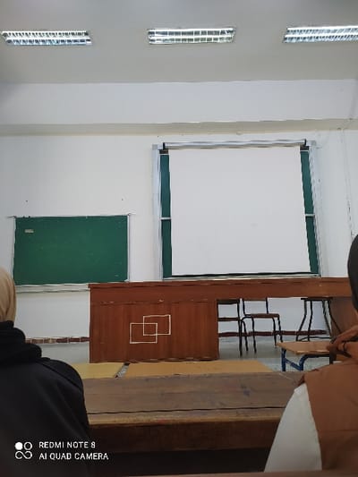 A classroom displays an overhead projector screen next to a wooden podium. A green chalkboard is positioned on the wall below the projector screen. Several chairs with metal frames are visible behind the podium. The room's interior is brightly lit, as indicated by the elongated windows on the left wall. The image also shows a partially obscured view of students in the foreground.