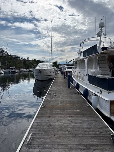The image depicts a marina in Nesøya, Norway. A wooden pier runs along the right side of the frame, adjacent to a body of water. Several yachts and sailboats are docked along the pier, some closer to the viewer than others. The boats vary in size and style, with a mix of white hulls and masts reaching towards the sky. On the opposite side of the water, there are trees and buildings visible on a hill. The sky is filled with clouds, reflected in the water's surface along with the boats and shoreline. The overall scene suggests a calm and peaceful atmosphere at the marina.
