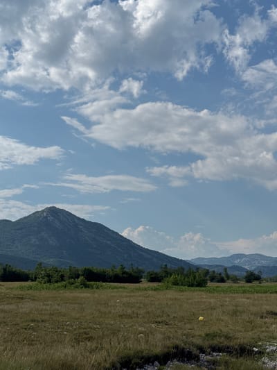The image depicts a landscape in the vicinity of Dragalj, Montenegro. A prominent, tree-covered mountain dominates the middle ground, its slopes receding into the distance. In the foreground, a dry, grassy field stretches out, dotted with scattered rocks. A single yellow object, possibly a piece of debris or litter, is visible in the field. The sky above is a mixture of blue and white clouds, with some rays of sunlight breaking through. The overall scene conveys a sense of natural, undeveloped terrain.