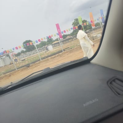 The image was taken in Wuro Ngangandi, Nigeria, showing a construction site with multiple colorful flags and banners hanging from a fence. A person dressed in a cream-colored robe is walking away from the camera on a dirt path. In the foreground, the dashboard of a vehicle is visible, with the word "AIRBAG" imprinted on it. The sky is overcast, and trees can be seen in the background. The construction site appears to be in an early stage, with piles of building materials present.