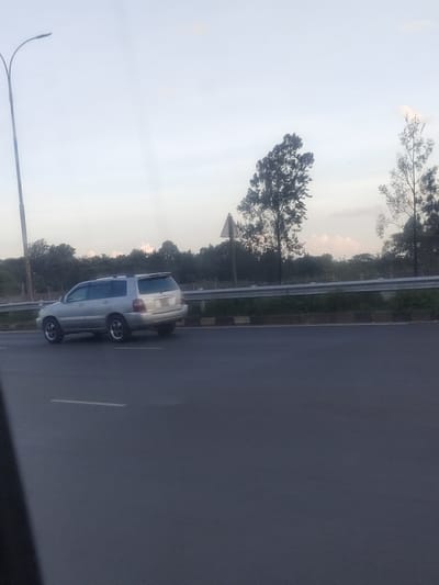The image displays an outdoor scene in Theta ward, Kenya, captured at either dawn or dusk, indicated by the pale, diffused light. A silver Toyota Highlander SUV is positioned in the mid-ground, traveling away from the viewer in the rightmost lane of a dark asphalt road. A white dashed lane marking is visible on the road surface in the foreground. To the far left, a tall, dark grey streetlamp with dual arms extends out of frame. A silver metal guardrail runs horizontally across the mid-ground, separating the road from an embankment. Below the guardrail is a low, two-toned concrete curb. Behind the guardrail, a triangular road sign on a pole is visible, and the area is densely vegetated with green ground cover and a line of dark green trees and bushes. A prominent, tall tree with a slight lean is situated to the right of the road sign. The background consists of a horizon line of dense trees beneath a pale blue-grey sky with subtle cloud formations. No individuals are present in the scene. A dark, blurry object partially obscures the lower-left corner, suggesting the image was captured from within a vehicle.