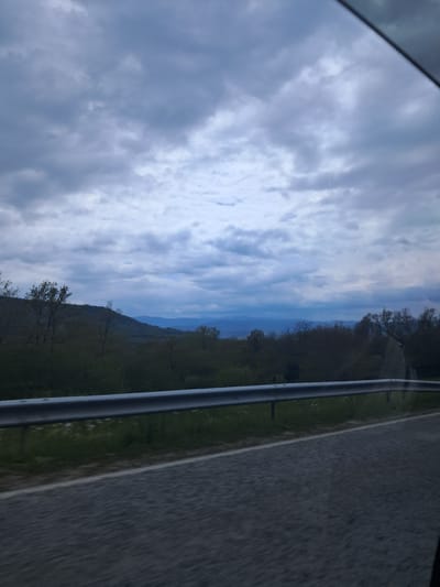 The image depicts a view from inside a vehicle, likely a car, showcasing a landscape in transit. The sky is filled with cloudy formations, occupying a significant portion of the frame. Adjacent to the sky is a view of a mountainous region, partially covered with trees and vegetation. Next to the green vegetation, there is an asphalt road.
The image was most likely taken in a mountainous area, but it's unclear if the picture location is in Хаджийско, Bulgaria.