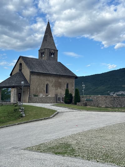 This media file is a photograph taken outdoors in Cavedago, Italy. The scene features a church with a tall, pointed steeple, likely made of stone. The church has a dark, sloped roof and arched windows. Adjacent to the church is a paved pathway, bordered by manicured green grass and stone walls. In the background, a steep, tree-covered mountain dominates the landscape under a bright blue sky with scattered white clouds. The lighting suggests it is daytime, possibly late morning or early afternoon. No people are visible in the photograph. The overall atmosphere is peaceful and serene, highlighting the architecture of the church and the natural beauty of the mountainous surroundings. The image is oriented vertically, with the sky and the top of the church steeple on the left side.