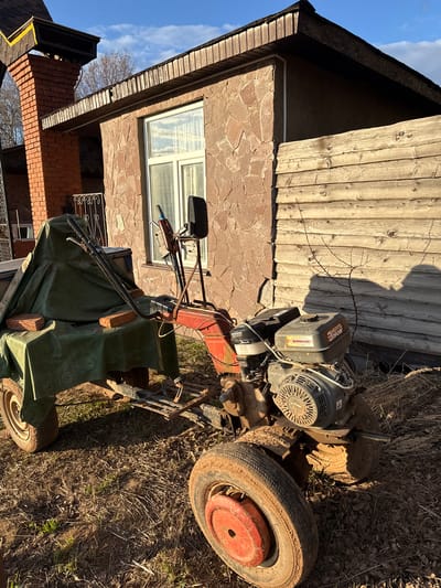 Outside a building in Galevo, Russia, a heavily soiled rototiller sits on dry grass and leaves. The building's facade is partially stone and partially wood. A small window is visible. The rototiller has a red and black engine and muddy tires. Part of the rototiller is covered with a green tarp secured with bricks. The sky is blue and sunny.