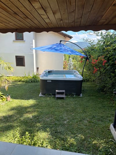 This is a medium-angle, full shot of a hot tub located in the garden of a house. The hot tub is dark gray with a white rim and appears to be filled with clear water. It is positioned on the grass, surrounded by greenery, and is partially covered by a blue umbrella with a patterned design. A small, wooden step leads up to the hot tub. To the left of the hot tub, a section of a white building is visible with a small, square window and a louvered vent. The garden features a lush green lawn, various plants, and a large bush with red flowers to the right of the hot tub. In the background, trees and rolling hills are visible under a partly cloudy sky. The scene is lit by natural sunlight, casting shadows on the grass. The image was taken outdoors, likely in a residential area. The presence of a hot tub and the surrounding vegetation suggests a garden or backyard setting, potentially in a suburban or rural area. Given the mention of Chavei, Bulgaria, it is likely a private residence in or near that city.