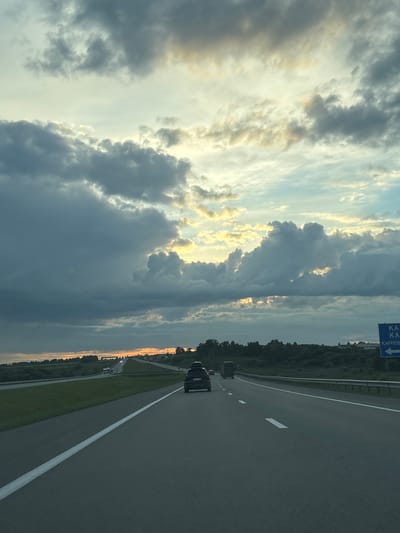 This is a photograph taken from a moving vehicle on a highway, likely in or near Kulaevo, Russia. The sky is dramatic, filled with a mix of dark, heavy clouds and patches of bright, golden light breaking through, suggesting either sunrise or sunset. The highway stretches ahead, with lanes marked by white lines. A dark-colored SUV is visible in the right lane, moving away from the camera, with a roof box on top. A truck is in the left lane, further ahead. The landscape on either side of the highway consists of green fields and a line of trees. A blue road sign is visible on the right side of the highway, with white text that appears to be in Cyrillic script and an arrow pointing left. The overall impression is one of a journey on a highway under a dynamic sky.