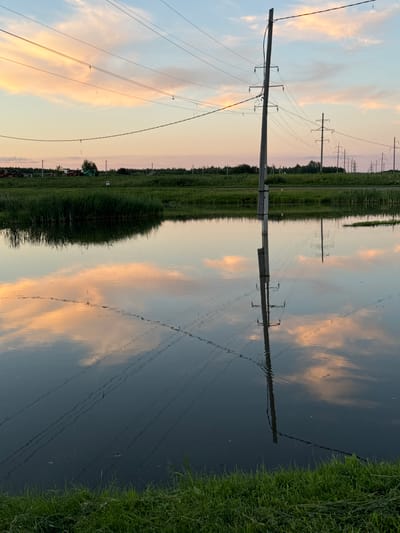 The media file displays a reflection of a rural landscape in water. The scene features a wooden utility pole with numerous power lines stretching across the frame. Beyond the pole, a line of green trees and vegetation is visible, with more utility poles and power lines in the distance. The sky is painted with soft hues of pink and orange clouds, suggesting a sunset or sunrise. The water's surface is calm, creating a clear reflection of the sky and the man-made structures. The presence of tall grass on the right side of the frame further emphasizes the natural setting. The image evokes a sense of tranquility and the quiet beauty of a rural evening. There is no visible text in the media file. The location is identified as Kommunar, Russia.