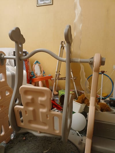 This is a horizontal, eye-level, close-up shot of a children's play structure and surrounding items in a room. The play structure, primarily made of beige and grey plastic and metal, occupies the foreground and mid-ground. It appears to be a multi-level climber or slide with various platforms, openings, and curved supports. On the left side, a framed picture hangs on a pale yellow textured wall, suggesting a domestic setting. To the right of the play structure, a fluffy grey rug is visible, indicating a floor covering.
In the background, more toys and household items are present, including stuffed animals, a blue hoop, and what looks like a white spherical lamp. A portion of a shelf with the word "HAPPY" is also visible. The scene is well-lit, suggesting it's daytime, and the overall impression is one of a playful, indoor environment. No people are visible in the frame. The focus is on the details and components of the play structure and the collection of items around it. The image is oriented such that the top of the image appears on the right, and the bottom on the left.