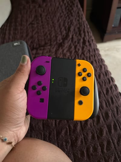 The image shows a Nintendo Switch console with a yellow top joy-con and a purple bottom joy-con. The console is being held in someone's hand, and a portion of their arm and bracelet are visible. The background features a brown textured surface that appears to be a couch or blanket. The image does not provide any information about the location, or city, or country it was taken in. Therefore, it's not possible to determine if the image was taken in La Aurora, Ecuador.
