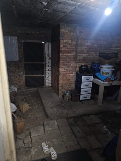 The image depicts an outdoor, cluttered storage area, possibly a basement or a backyard shed in Daskotna, Bulgaria. The scene is dimly lit by a single overhead light fixture. The walls are constructed of red brick.

In the background, a door with a screen is visible, with a white plastic-covered object to its left. To the right of the door, a tall, narrow wooden cabinet with four drawers is placed. On top of a low table next to the cabinet, there are two large pots, one with a lid and a fruit pattern, and a blue plastic barrel. Cardboard boxes are stacked behind the table.

The ground is uneven and covered with debris, including scattered pieces of wood and what appears to be concrete or stone slabs. In the foreground, a pair of white sandals lies on the ground.