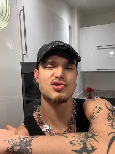 A male individual is depicted in a close-up, chest-up self-portrait within a modern kitchen setting in Asnières-sur-Seine, France. He wears a distressed black baseball cap, a dark sleeveless top, and a prominent silver-toned chain necklace with spiky elements. His left ear features multiple silver-toned earrings, including a large hoop. Dark, intricate tattoos cover both of his visible arms. He has a goatee and mustache, and his facial expression shows narrowed eyes and pursed lips. The background consists of white, high-gloss flat-panel kitchen cabinetry with metal handles. An integrated dark-glass appliance, likely an oven, is visible on the left. On the right, a light-colored, speckled countertop holds a small red vase containing dried red flowers. A ribbed glass light fixture is partially visible in the upper left quadrant.