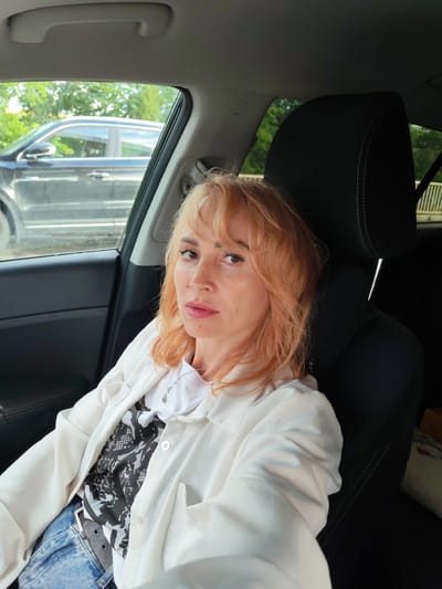 A medium shot taken from the interior of a car shows a woman with ginger hair sitting in the passenger seat. She is looking directly at the camera with a neutral expression. The woman is wearing a white collared shirt over a black and white patterned t-shirt, blue jeans, and a black belt. A silver necklace with a small pendant is visible. The car's interior, including the black headrest with green stitching, is visible. Through the car window, another dark-colored vehicle is seen in motion, along with blurred greenery and a glimpse of a fence. The scene is outdoors, suggesting a setting that could be in or around Staraya Yum'ya, Russia, given the time of year likely corresponding to warmer weather due to the greenery.