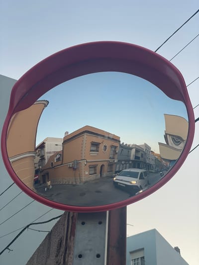 The image shows a convex mirror, likely a traffic mirror, with a red plastic frame, mounted on a metal post. The mirror reflects a street scene in La Línea de la Concepción, Spain. The reflection reveals a street lined with buildings, some of which have a light orange facade and decorative elements. A white van is visible driving on the street, with its headlights on. Further down the street, other vehicles and buildings are reflected. The sky in the reflection is a clear blue. In the foreground, outside the mirror, a rough stone surface and part of the metal post are visible, along with electrical wires.