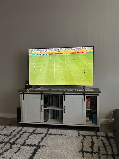 A wall-mounted television displays a soccer game in progress on a green field. Adjacent to the TV, a white cabinet with rustic hardware sits. The cabinet features two closed compartments with doors and an open shelf filled with books. A gray and white rug with a geometric pattern lies on the floor near the cabinet. The background includes a plain gray wall.