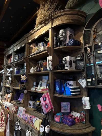 A tiered wooden display case showcasing various items, predominantly Harry Potter merchandise. The top shelf features masks, a Deathly Hallows emblem, and miscellaneous accessories. Subsequent shelves hold character figurines, stationary items with Luna Lovegood branding, and other related products like journals, and tote bags. A broom is positioned to the left of the case.