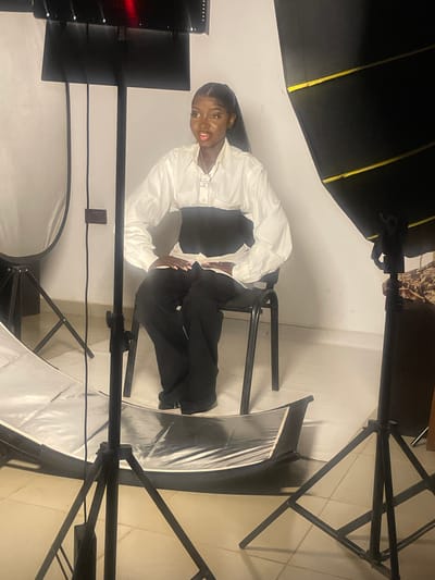 In South Ropp, Nigeria, a young Black woman with her hair pulled back in a ponytail sits in a black office chair in what appears to be a photography studio. She is wearing a white button-down shirt with a black corset-like garment cinched around her waist, and black wide-leg trousers. She is adorned with a necklace. Her hands are resting on her lap, and she is looking slightly to her left with her mouth open. The studio is equipped with professional lighting equipment, including a large softbox to the right and a reflector with a stand to the left. There is also a large overhead light fixture. The walls are white, and the floor is tiled. The overall scene suggests a posed photo session.