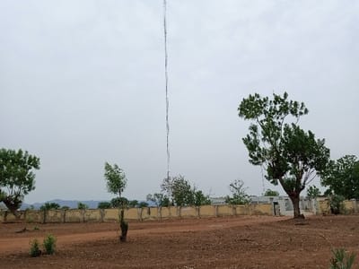 The image shows a wide, open field with a tall metal antenna in the center, extending from the ground into a cloudy sky. The antenna is the focal point, with the rest of the scene receding away from it. On the left side of the image, a small tree stands close to the camera, while a larger tree stands on the right side of the image. The scene is surrounded by a low wall and several trees, some bare and some leafy. The field is dry and dusty. There are no people or animals in the image. The coordinates (latitude: 9.189, longitude: 12.523) place the scene in Rumde Waziri, Nigeria. The image appears to be taken from a moderate distance away. The scene appears to be a rural or suburban area. The time of day appears to be late morning or early afternoon, based on the angle of the sun and the shadows cast by the trees.