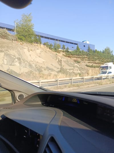 This is a first-person perspective image taken from inside a vehicle, likely while driving on a highway. The dashboard is visible in the foreground, displaying a digital readout of "86" and other indicators. Through the windshield and side window, a dry, rocky hillside is prominent, with sparse vegetation including bushes and small trees.

Dominating the upper part of the image is a large, modern-looking building with a blue roof and facade, featuring numerous windows. A small turret with a white and blue patterned roof is visible on the right side of the building. Evergreen trees are planted in front of and around the building.

A highway guardrail runs parallel to the road, separating the lanes of traffic. On the highway, a white truck is visible, as well as a white car. The sky is clear and blue, suggesting a sunny day. The lighting indicates it is either morning or late afternoon. The overall scene depicts a landscape that is somewhat arid and hilly, with modern infrastructure. The image appears to have been taken in Yahşihan, Turkey, based on the provided location context. There is no visible text other than the digital display in the car.