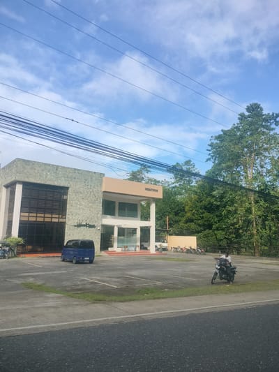 The image captures a front view of the Wood House Crematorium in Kidapawan City, Philippines. The building, constructed with a combination of stone and modern architectural elements, features a large glass facade on the left and a lighter-colored section with glass windows on the right. A blue minivan is parked in front of the building. The area is a parking lot with visible parking lines and some patches of grass. In the background, a line of motorcycles is parked, and a person is seen riding a motorcycle on the right side of the image. Behind the building, dense green trees form a natural backdrop, and a clear blue sky with scattered clouds dominates the upper portion of the scene, with numerous electrical wires crisscrossing overhead. The ground in the foreground is an asphalt road.