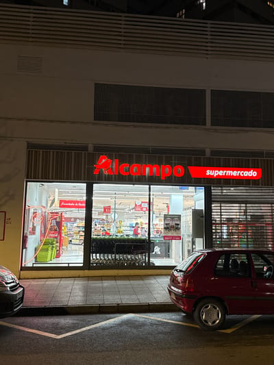 The image shows the exterior of an Alcampo supermarket at night in Oviedo, Spain. The illuminated "Alcampo Supermercado" sign is vertically aligned along the building's facade. Inside, the supermarket is brightly lit, revealing aisles of products, shelves, and promotional displays. Shopping carts are visible near the entrance. A red compact car is parked partially blocking the entrance, with its rear end angled towards the building. A second vehicle is partially visible at the bottom edge of the picture. The building's facade consists of gray concrete walls with rectangular grilles and vertical vents. The sidewalk next to the building features a painted yellow line and a dark grey asphalt surface.