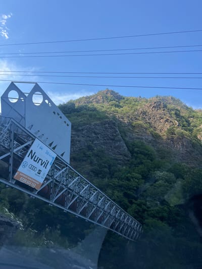 An angled, eye-level shot captures a section of a metal bridge structure with a large white sign attached to it. The sign displays "Nurvil" in large, black lettering, with smaller text above and below. To the left of "Nurvil" is an icon of a car and a truck, and to the right, "36 km" is written on a white banner. Beneath "Nurvil" are logos for Audi, Volkswagen, Seat, and Skoda. A smaller orange banner at the bottom of the sign reads "ASISTENȚĂ TEHNICĂ NON STOP: 0720 112 112". The bridge appears to be suspended over a forested valley, with a steep, rocky mountain covered in dense green foliage rising in the background. The sky is a clear, bright blue with a few wispy white clouds. Power lines are visible against the sky above the mountain. The overall scene suggests a scenic route passing through a mountainous region near Brezoi, Romania.