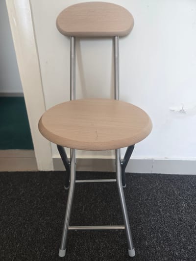 The image shows a close-up, rotated view of a wooden stool with silver metal legs. The stool has two oval-shaped wooden seats, one in the foreground and another behind it. The legs are thin, cylindrical metal rods that extend downwards from the seats. A dark grey carpet is visible on the right side of the image, and a white wall with peeling paint is on the left. The lighting suggests it is daytime, and the indoor setting with carpet and a wall implies a domestic or commercial space. There is no text visible in the image, and no people or discernible activity are depicted. The focus is solely on the stool and its immediate surroundings. The location context provided is Deira, United Arab Emirates, but this cannot be visually confirmed from the image itself.