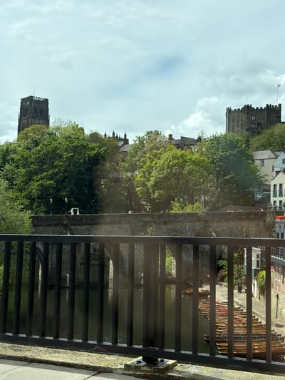 The photograph is taken from a bridge in Durham, United Kingdom. A dark metal railing dominates the foreground. Beyond the railing, the River Wear is visible, with arches of a stone bridge reflecting in the water. Lush green trees line the riverbank, obscuring some of the view. In the background, the Durham Cathedral and Durham Castle are visible, landmarks of the city. Other buildings, some residential, are nestled among the trees. The sky is overcast with scattered clouds. The scene captures a blend of architectural heritage and natural landscape typical of Durham.