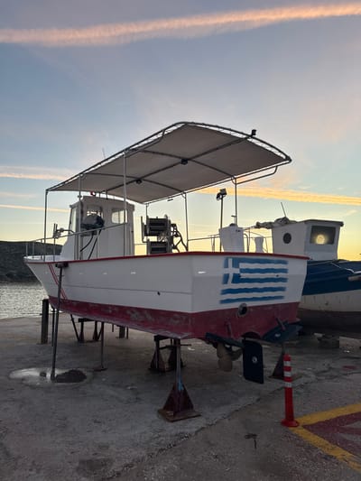 Two fishing boats sit on a concrete platform. The closer boat is predominantly white with a red stern and features the Greek flag. A canopy covers part of the boat. A propeller is visible at the back. The boat is supported by metal stands. The second boat, mostly blue, is partially visible behind the first. The sky in the background is a light blue with a hint of yellow and streaks of contrails. A red and white bollard sits beside the boat.