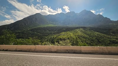 This image depicts a panoramic view of a mountainous landscape under a bright, sunny sky with scattered clouds. The mountains are covered in lush green vegetation, with dense forests and smaller bushes dotting the slopes. In the foreground, a paved road is visible, separated from the landscape by a concrete barrier. The sun's rays are shining brightly, creating a slightly hazy effect in the distance and highlighting the textures of the mountains. There are no people or man-made structures other than the road and barrier visible in the scene. The location is identified as the city of Zintsar, Russia.