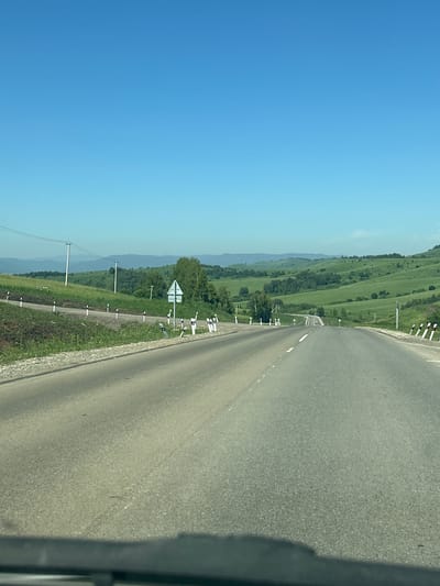 A paved road curves through rolling green hills under a clear blue sky. The road is lined with white and black markers, and on the left side, there are power lines supported by poles. In the distance, the road leads towards more hills, with a few vehicles visible. The foreground is obscured by a dark shadow, possibly from a vehicle. The scene appears to be in a rural or semi-rural area, consistent with a location like Verkh-Aya, Russia.