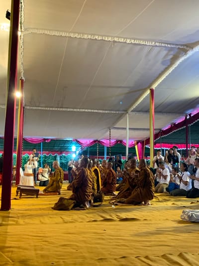 The image shows a group of Buddhist monks in saffron robes kneeling and praying in a large, white tent. They are facing the left of the frame, and their heads are bowed. The floor is covered with a golden-colored carpet. The tent is supported by red and yellow poles, and there is a red curtain lining the back wall. There is a row of people in the background, mostly women, who are observing the monks. The event appears to be taking place in an open space, with trees and greenery visible in the background. This event is most likely taking place at the Vihara Avalokitesvara temple in Mungkid, Indonesia, which is located at latitude -7.605 and longitude 110.23.