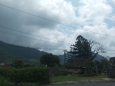 A serene landscape unfolds beneath a sky of clouds ranging from light to dark. Lush green mountains form a backdrop, partially obscured by trees. Wires run across the frame, adding a human-made element to the natural scene. In the foreground, a quaint building with a red-tiled roof peeks through foliage. A utility pole stands tall beside it, connecting civilization with the tranquility of nature. A hedge and partial fence define the boundary.