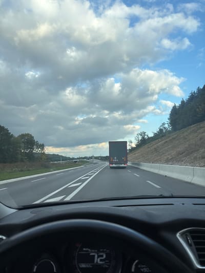 The image depicts a daytime view from inside a vehicle traveling on a multi-lane highway in the vicinity of Lièpvre, France. In the foreground, the upper portion of a black steering wheel and parts of a dashboard are visible, including an instrument cluster displaying "72," likely indicating speed in km/h.

The midground shows a paved highway stretching into the distance. The vehicle from which the image is taken is in the right lane, following a black semi-trailer truck. The truck's rear features red reflective markers and two circular "00" signs, along with a "P" sign. The highway has distinct white dashed and solid lane markings. To the right of the highway, a concrete barrier separates the road from a steep, partially vegetated embankment leading to a dense forest of green trees. To the left, a grassy verge borders the road, followed by a line of trees, and in the far distance, a bridge or overpass structure is discernible.

The background consists of a partly cloudy sky, featuring a mix of white and grey cumulus clouds, with intermittent patches of clear blue sky, predominantly towards the upper right. The overall lighting suggests ambient daylight. No individuals are visible in the scene.