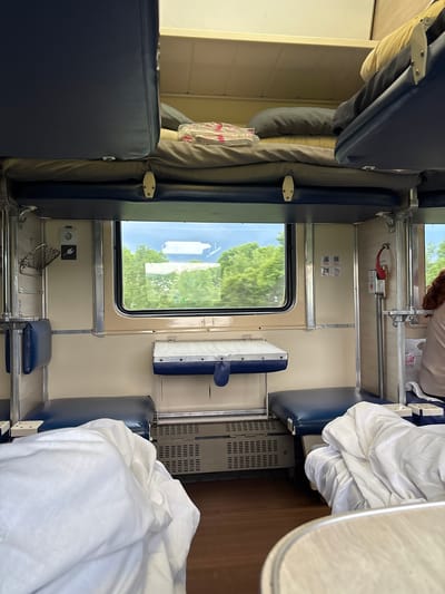 The image shows the interior of a train compartment, likely a sleeper car. The compartment features bunk beds with blue upholstery and white linens. There is a window with a view of green trees and a cloudy sky, indicating it is daytime and the train is in motion. The interior walls are beige, with silver accents and various fixtures, including an electrical outlet, a small basket, and a fire extinguisher. The floor appears to be dark wood. No people are clearly visible in the main area of the compartment, though a person with red hair can be seen in the background on the right. The location "Raz"Yezd 9208 Kilometr, Russia" is a point on a railway line and not a city or town, suggesting a precise geographical reference within Russia's railway network rather than a typical urban location.